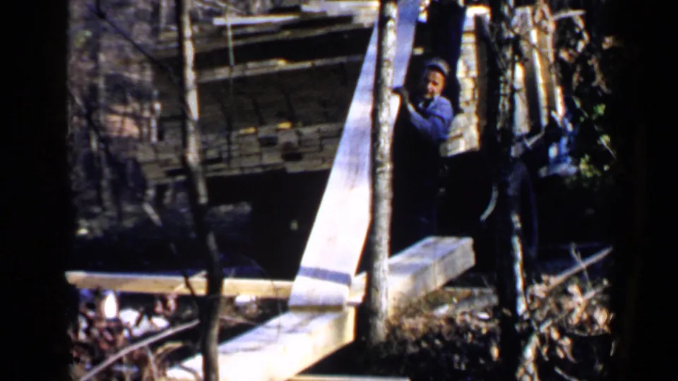 1957 Birmingham-area frame with a man in work clothes handling a long pale plank beside a truck in the woods.