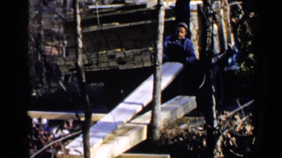 1957 Birmingham-area home-movie frame showing the man bent over as a long board projects across the wooded work area.