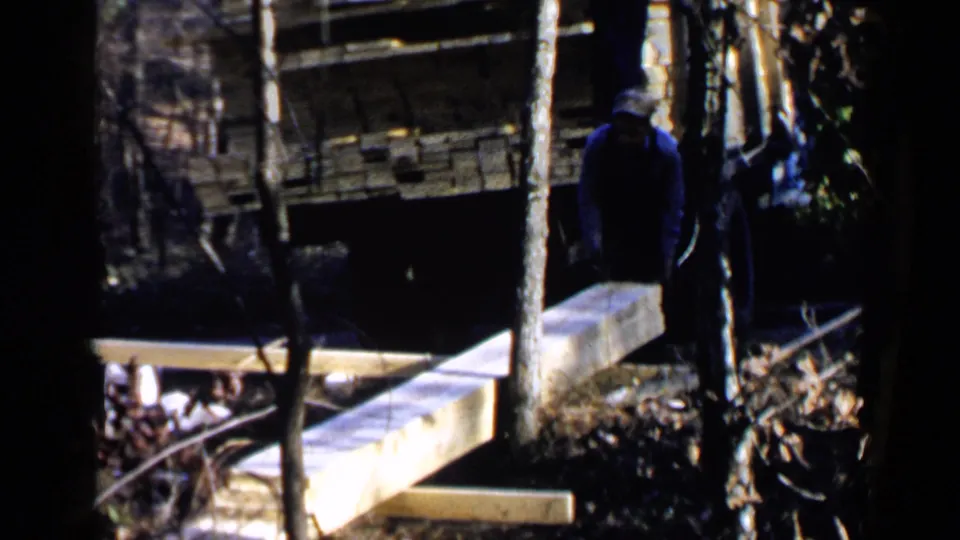 1957 Birmingham-area 8mm frame with the man turned away beside long boards, trees, and a truck or trailer bed.