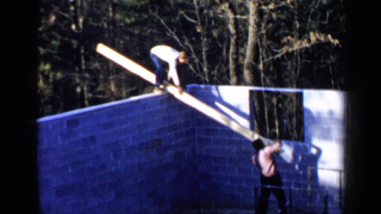 Two men position a long wooden beam on a cinder block wall while another stands below.