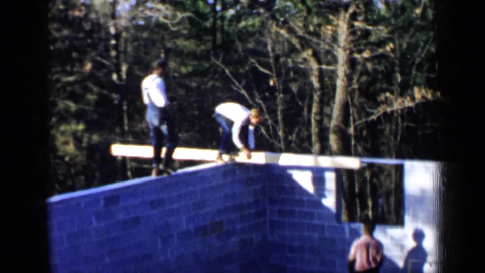 Adult men in overalls and light shirts work on a cinder block foundation, with a long beam spanning the wall in a wooded setting.
