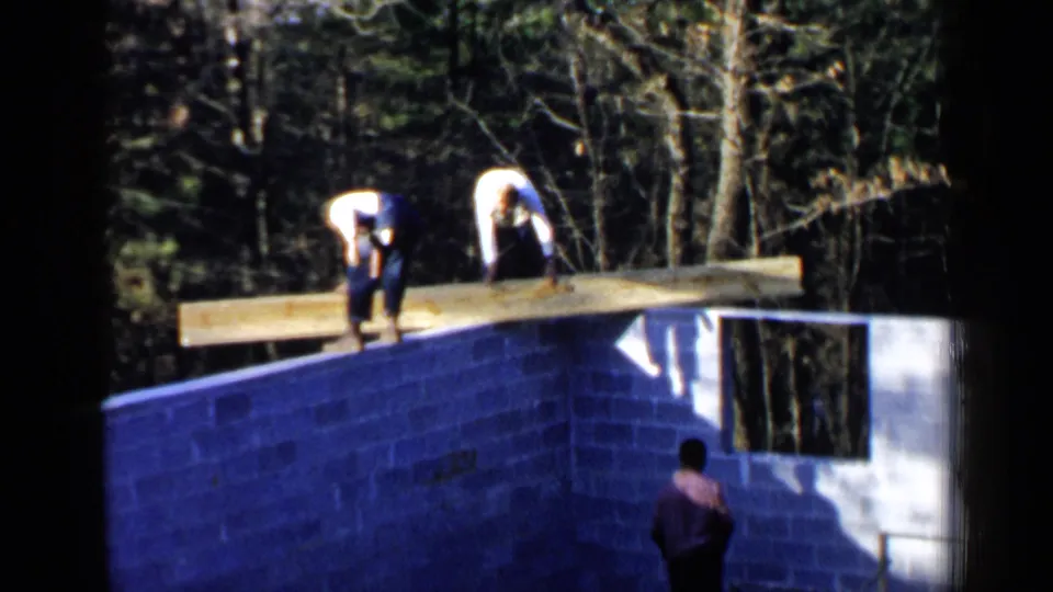 At the end of the clip, two workers bend over the lumber on the cinder block wall as a third man watches from below in daylight.