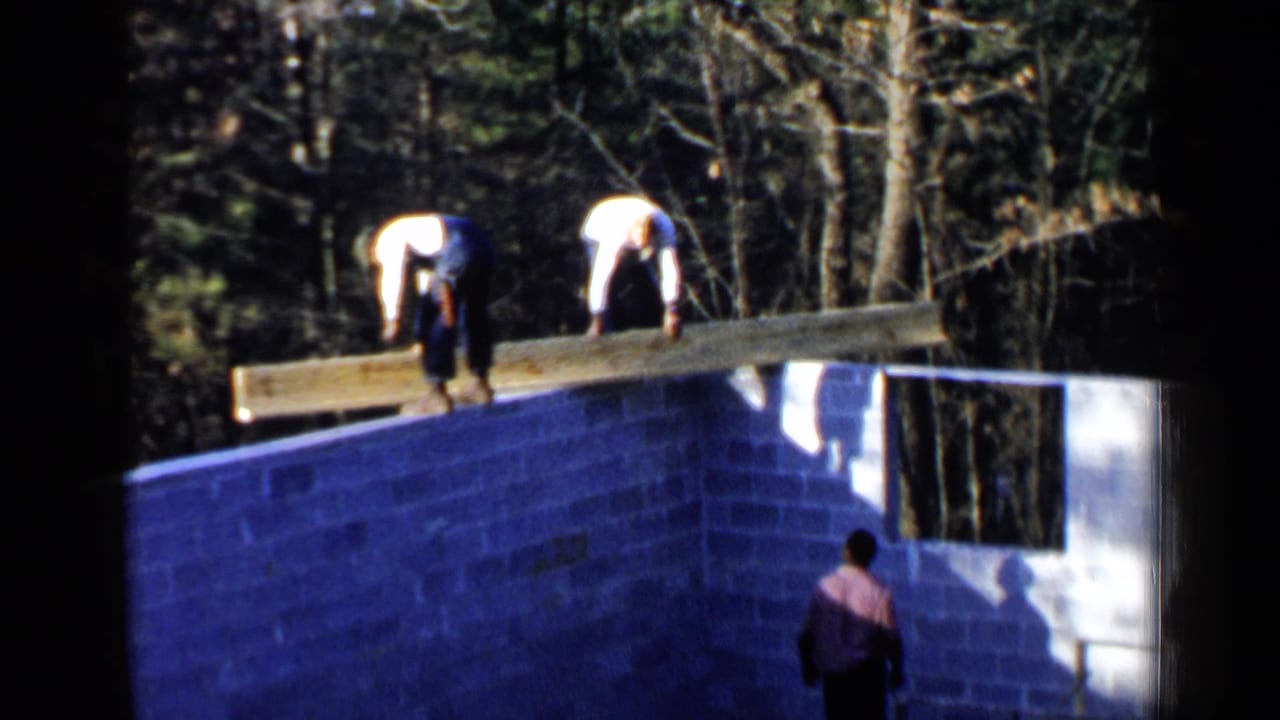 1957 Birmingham home-movie frame showing men positioning a long wooden beam across a cinder block foundation in a wooded setting.