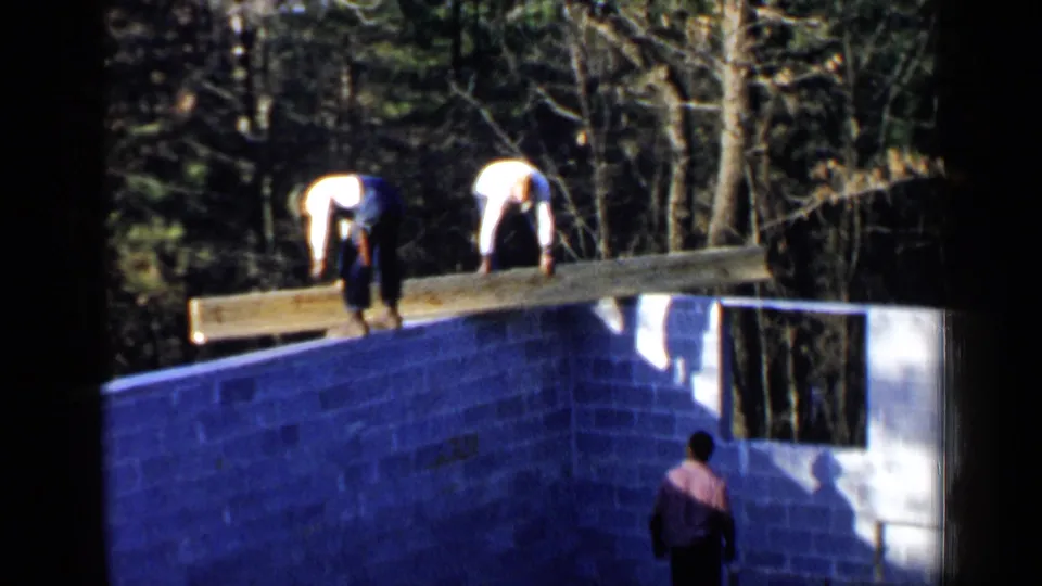 1957 Birmingham home-movie frame showing men positioning a long wooden beam across a cinder block foundation in a wooded setting.