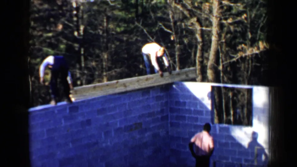 Birmingham 1957 frame with men bending over a long beam atop an unfinished cinder block wall, filmed from a home-movie viewpoint.