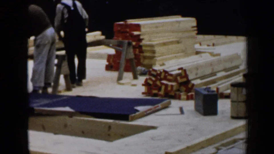 Two workers are visible on a 1957 Birmingham construction site near stacked lumber, red brackets, and a sawhorse.
