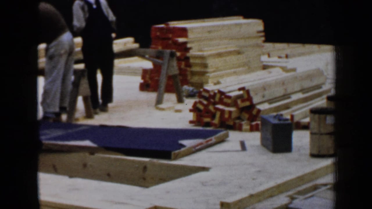 1957 Birmingham construction scene with workers beside lumber stacks, boxed materials, sawhorses, and a blue tarp on the ground.