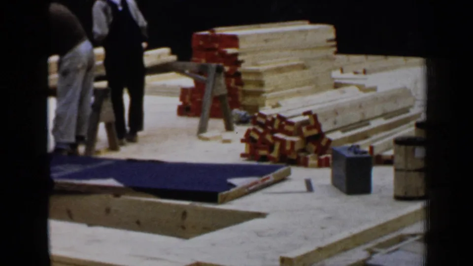 1957 Birmingham construction scene with workers beside lumber stacks, boxed materials, sawhorses, and a blue tarp on the ground.