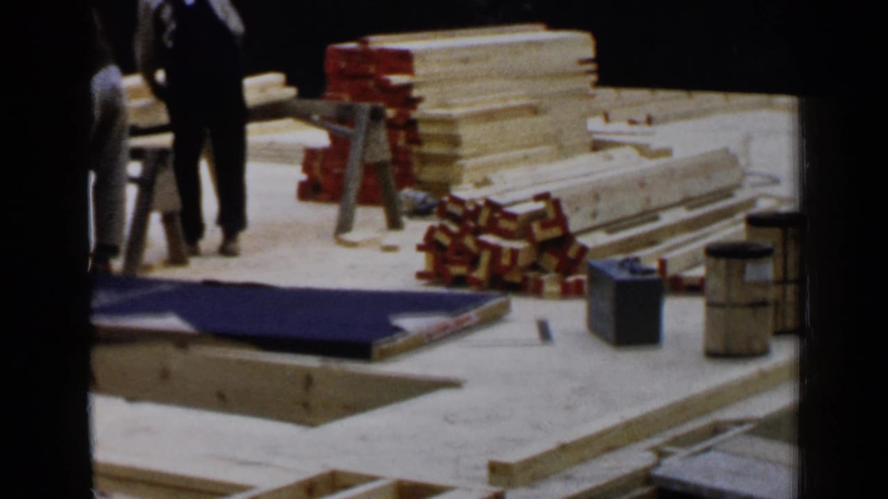 Workers and construction supplies appear beside stacked lumber and a blue tarp.