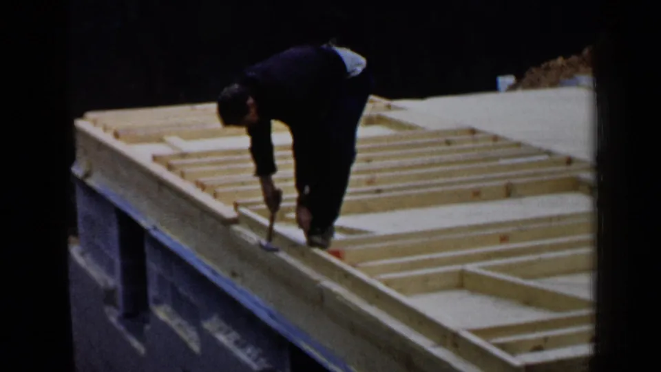 A worker in 1957 Birmingham bends over open wood framing, hammering joists into place on an unfinished house structure.