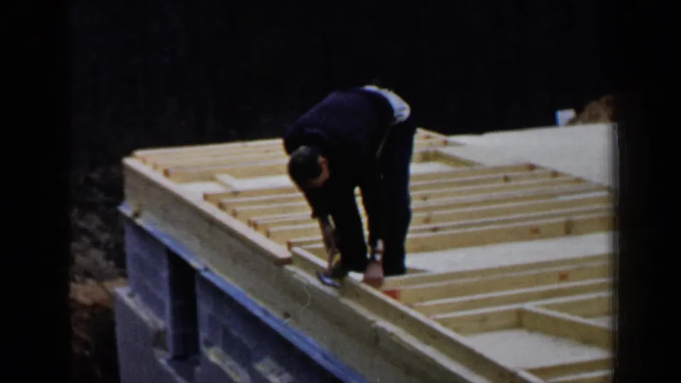 A worker continues hammering or positioning wooden joists on an unfinished 1957 Birmingham house frame.