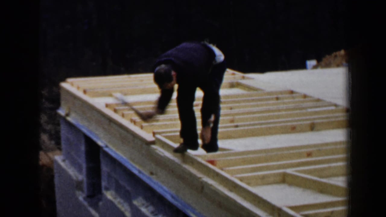 A worker reaches across pale wooden roof framing on a sloped or elevated structure, with dark trees behind the construction area.