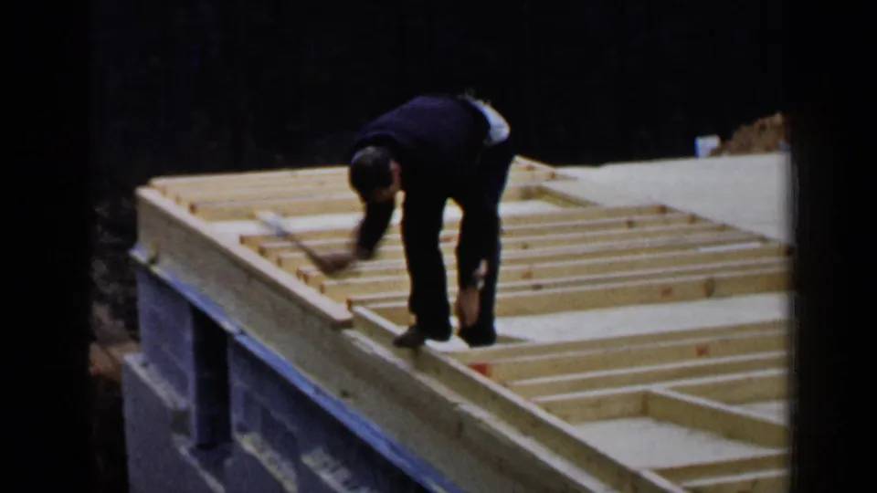 A worker reaches across pale wooden roof framing on a sloped or elevated structure, with dark trees behind the construction area.