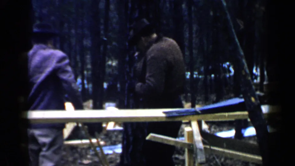 Two adult men stand beside a long plank on supports in a wooded construction area, continuing the home-movie record of building work.