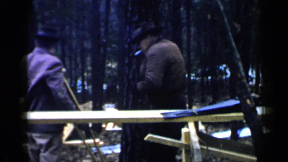 1957 Birmingham-area 8mm frame showing two adults near wooden planks and supports among tall trees.