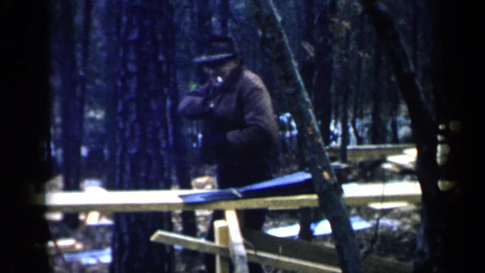 A man in a dark hat and coat stands near a wooden work structure in the Birmingham 1957 home-movie scene.