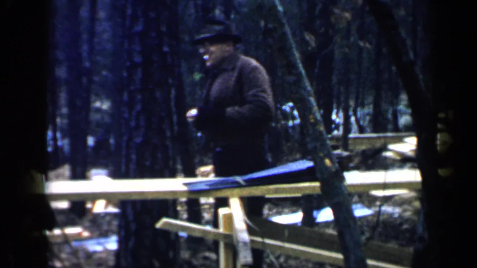 The man appears to hold a small object after reaching into his coat, with wooden planks and trees behind him.