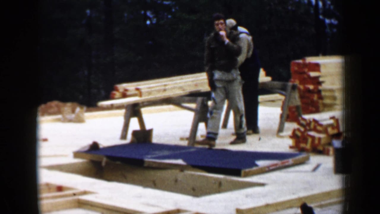 Two men stand near lumber on sawhorses at an outdoor house construction site.