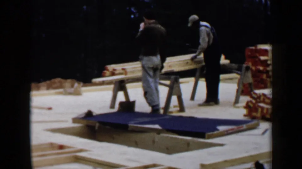Two adult men work with long boards on sawhorses at a wooded 1957 Birmingham residential building site.