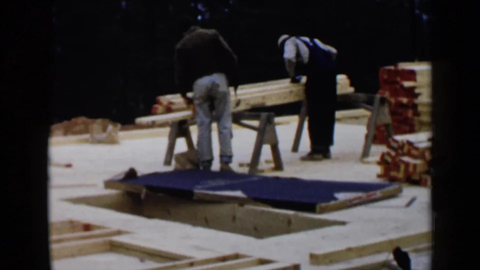 Home-movie view of two men working over boards and materials on a wooden house platform in Birmingham, Alabama, in 1957.