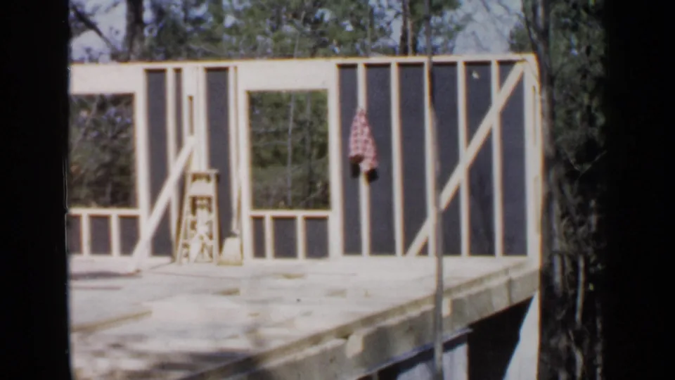 Wooden wall framing stands on a raised platform at the Birmingham house construction site, with trees visible behind the structure.