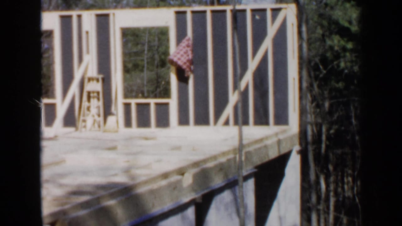 Unfinished wooden house frame with ladder, broom, checked cloth, and trees beyond.