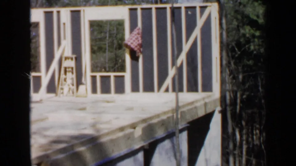 1957 Birmingham home-movie frame showing an unfinished wooden house structure with ladder, broom, checked cloth, and wooded surroundings.