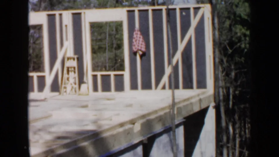 The 1957 Birmingham footage frames an unfinished second-floor house area with exposed studs, tools, checked cloth, and trees beyond.