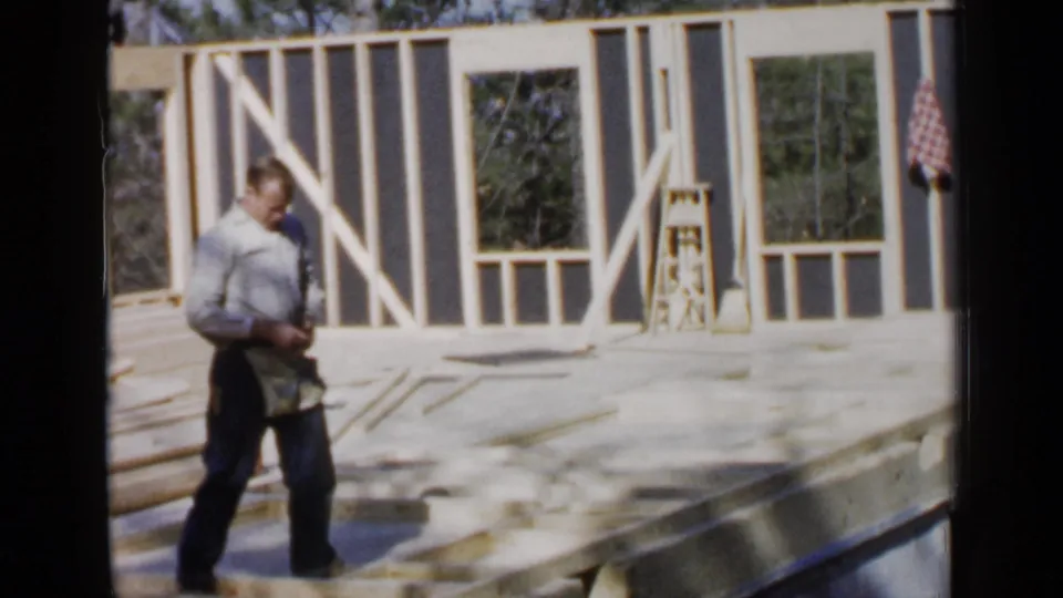 In the 1957 Birmingham home movie, an adult man crosses the unfinished second floor of a framed house with tools nearby.