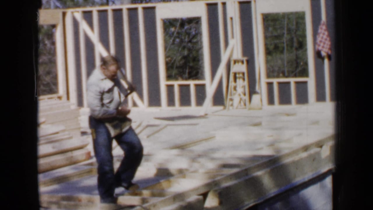 Man stands on an unfinished wooden house frame holding a tool near framed wall openings.