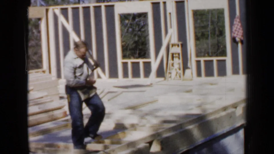 In 1957 Birmingham, Alabama footage, a man stands on a sunlit wooden house frame with wall studs and window openings behind him.