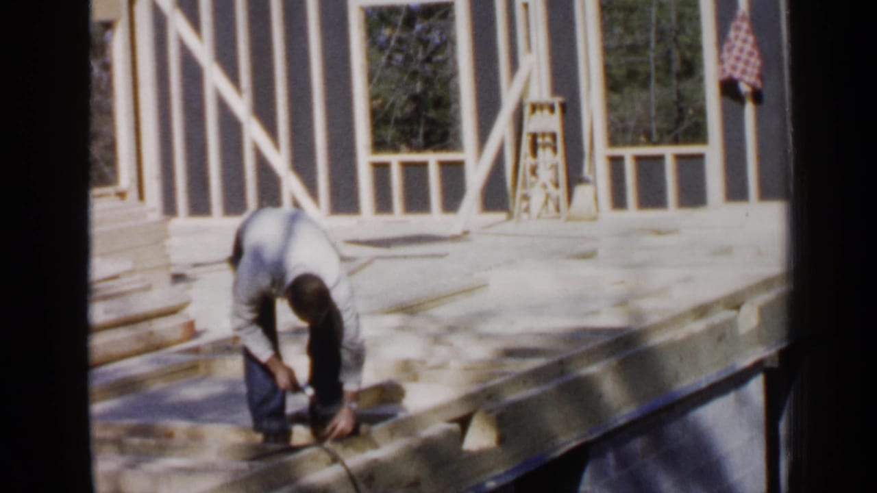 Man bends over the edge of an unfinished wooden floor while working with a tool.