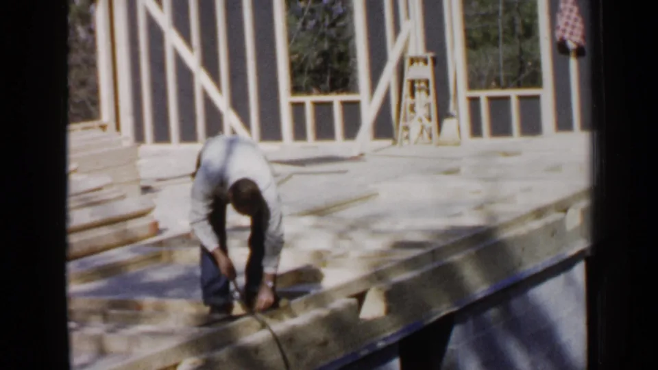 A 1957 Birmingham home-movie still shows the man leaning down over unfinished wood framing, with sunlit studs and open window bays in the background.