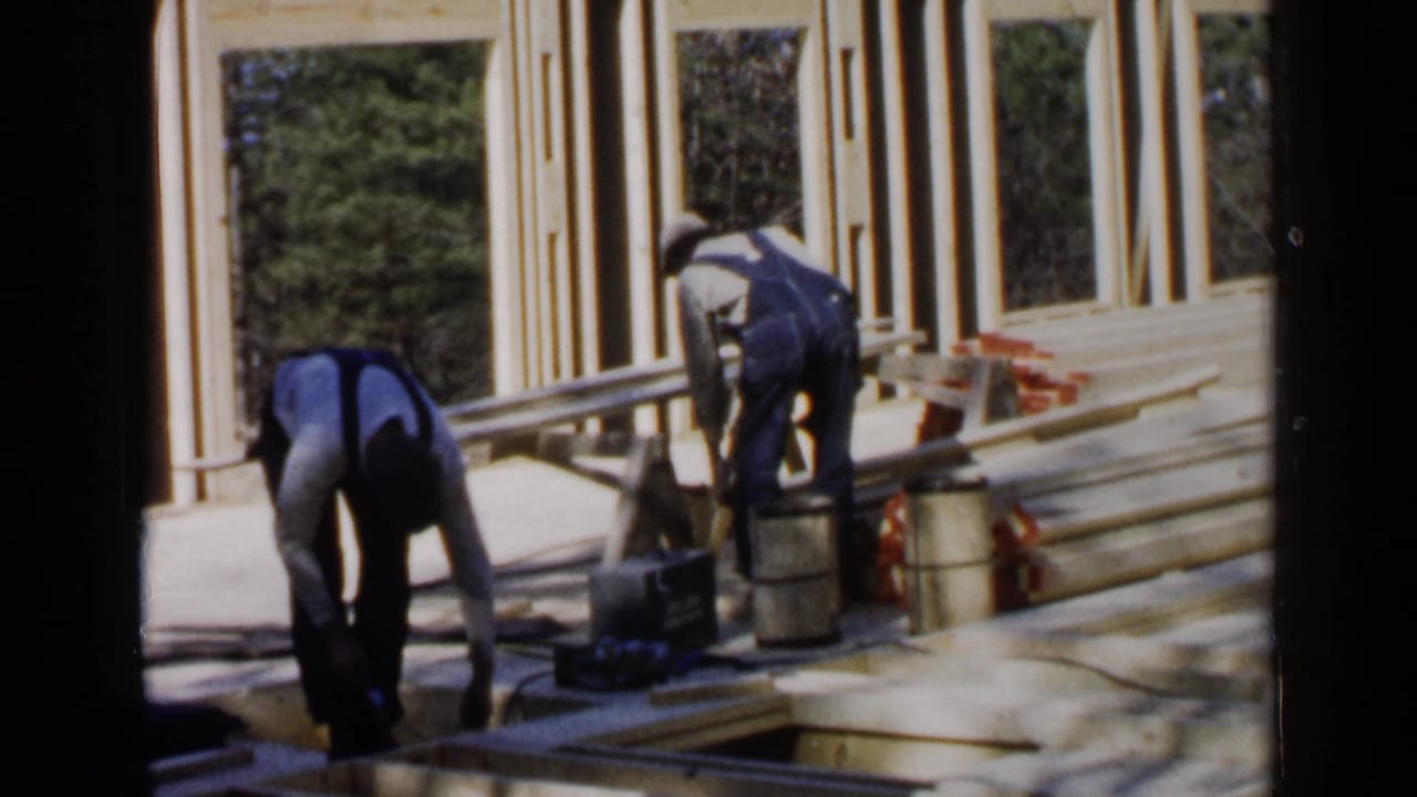 Two men bend over lumber beside a partially framed wooden structure.