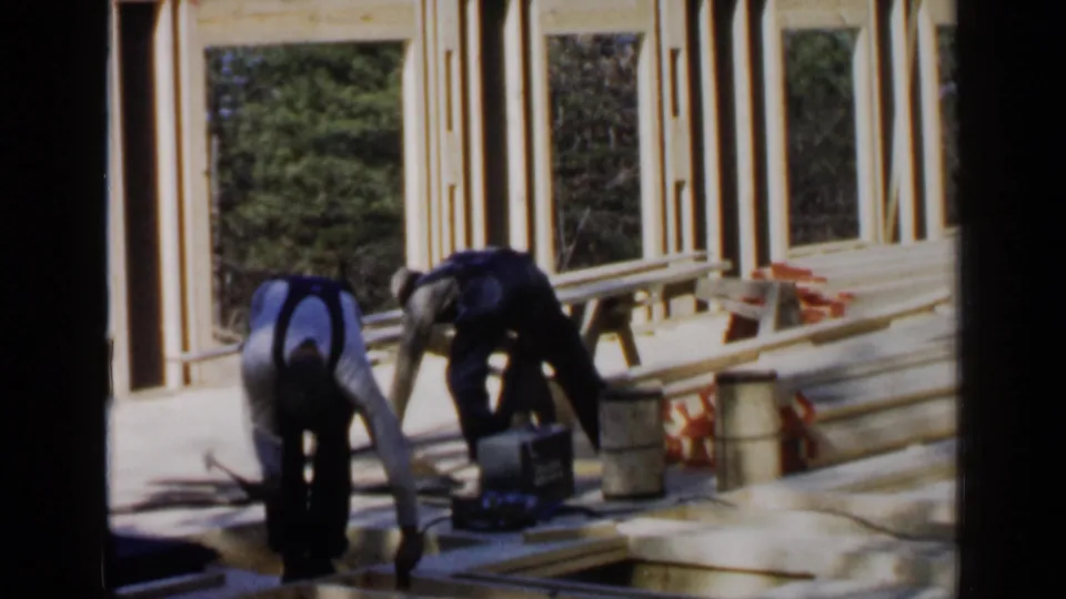 At the 1957 Birmingham construction site, two workers handle lumber near a wall frame with barrels and tools nearby.