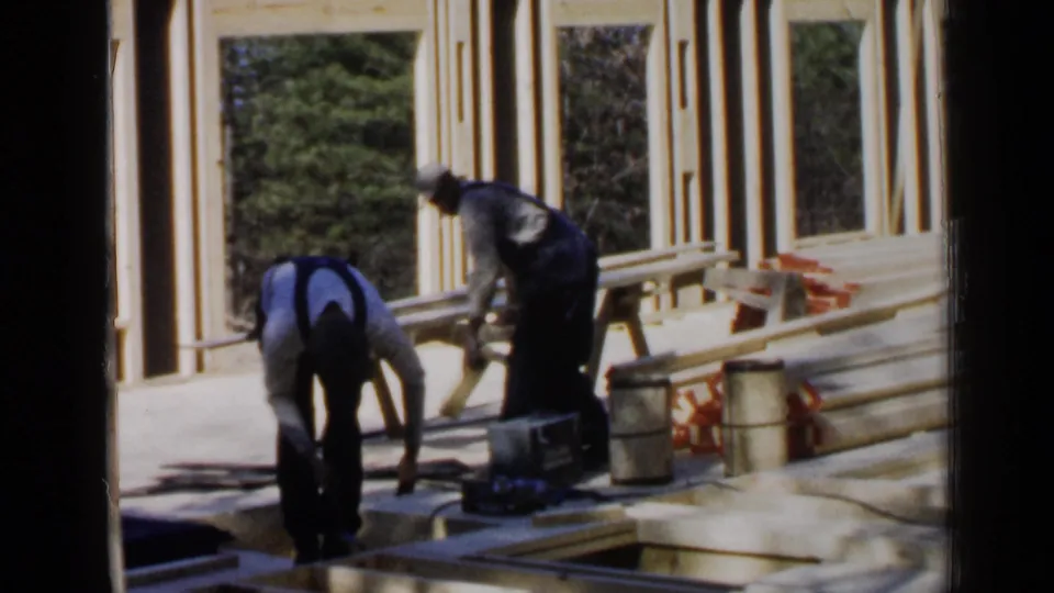 One worker stands while another bends over boards beside framed openings at a 1957 Birmingham building site.
