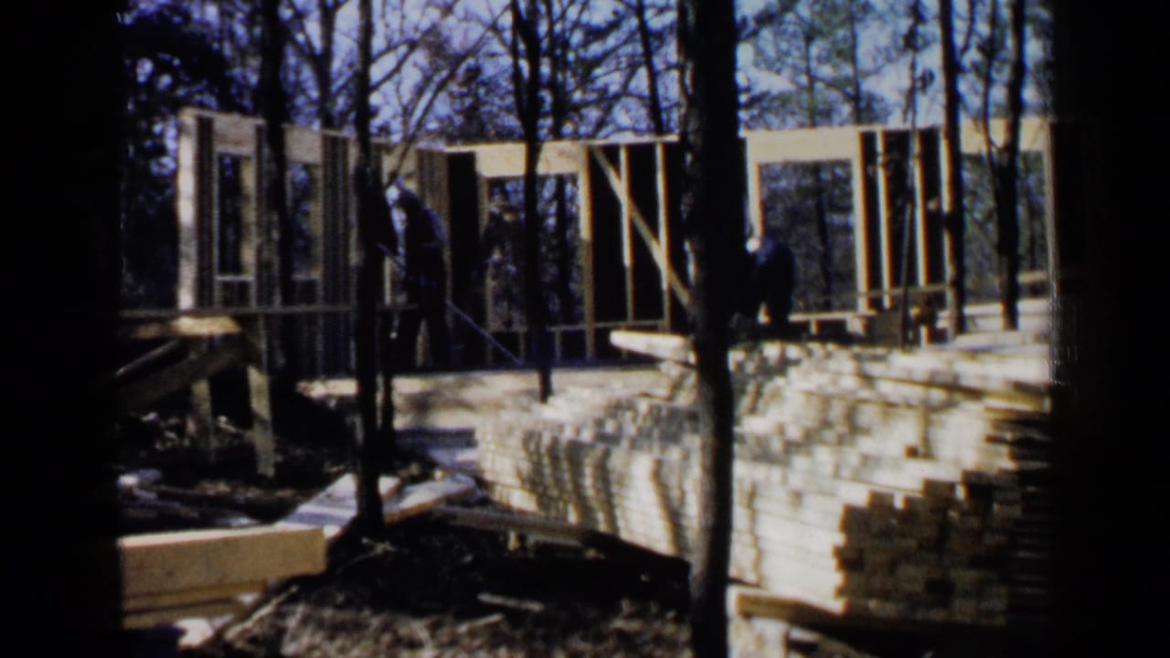 Wooden house frame under construction among trees with lumber stacked in front.