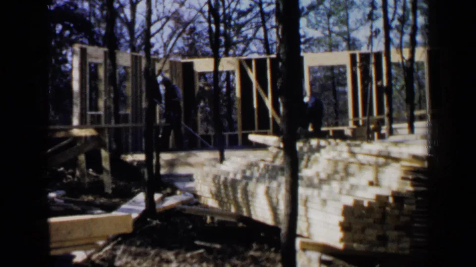 1957 Birmingham-area 8mm frame showing workers near a partially built wooden house frame in a wooded setting.
