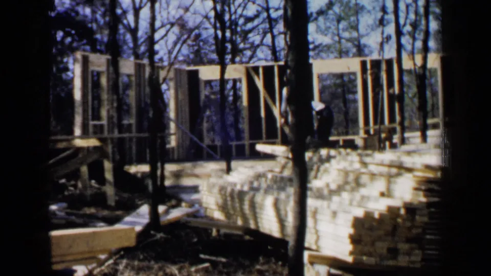 Wooden wall framing and stacks of lumber appear through trees during 1957 Birmingham-area house construction footage.