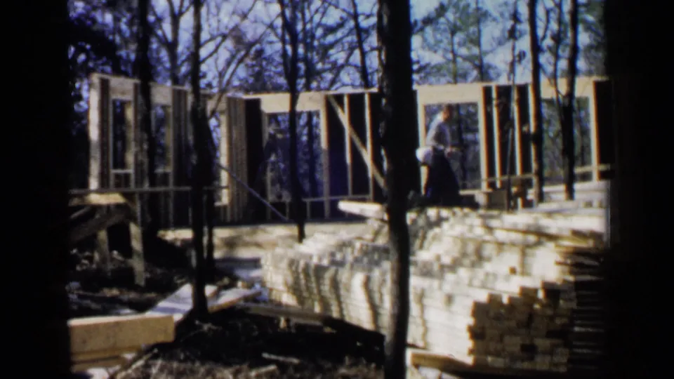 Handheld 8mm view of a wooded Birmingham construction site with workers beside upright house framing and stacked boards.