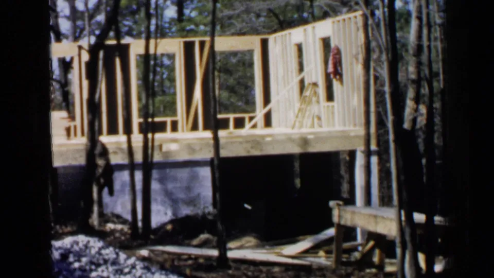 A raised wooden house frame in a wooded Birmingham setting, with a worker below and construction materials nearby.