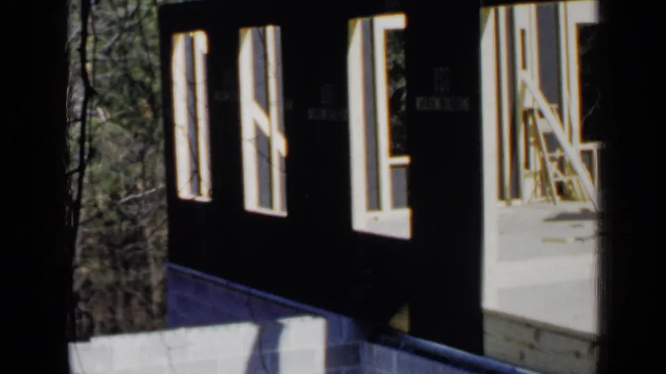 1957 Birmingham 8mm frame showing an unfinished house wall with open window openings, new lumber, and trees beyond.