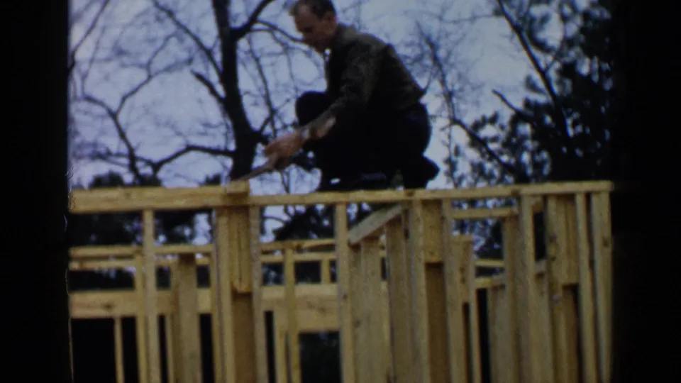 A middle-aged man in 1957 Birmingham crouches on exposed wooden framing and works with a hammer against a backdrop of bare trees.