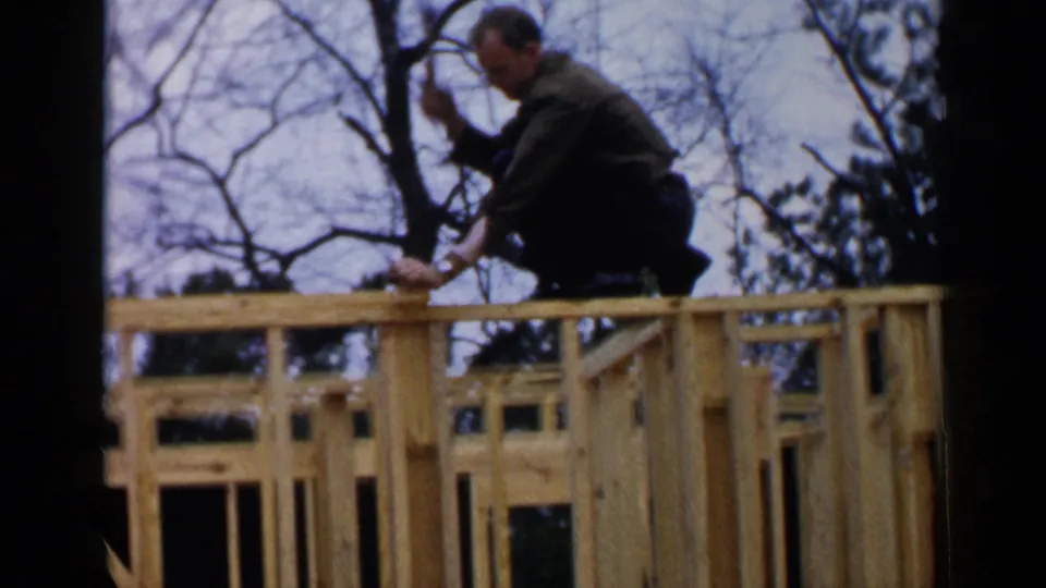 1957 Birmingham frame showing a man crouched on new house framing, hammering wood with bare trees and blue sky behind him.