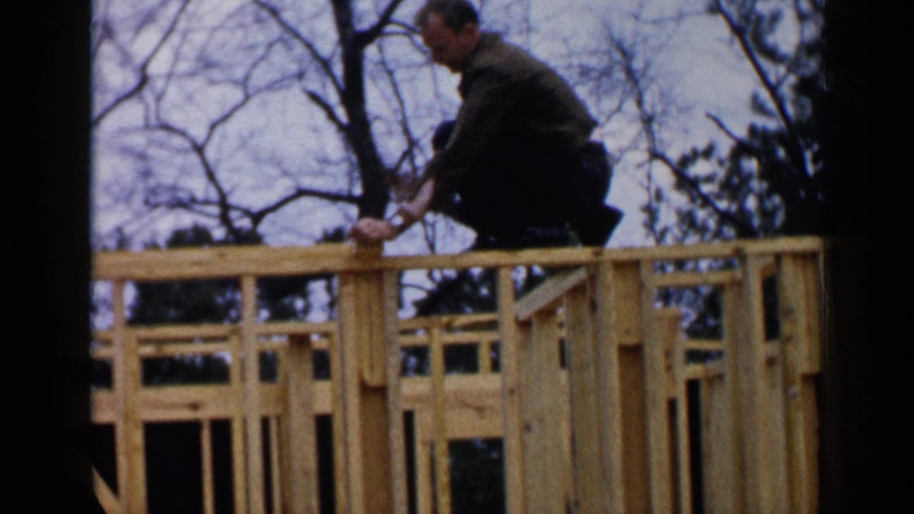 Man in dark clothing crouches on the upper wooden frame of a house under construction.