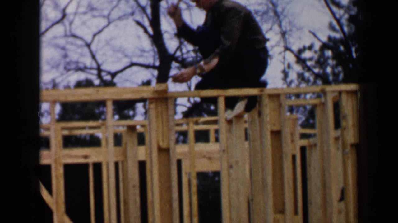 Worker leans over unfinished wooden framing with trees visible behind him.