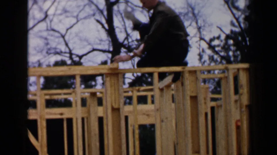 8mm home-movie still of a man positioned on the upper framing of a house under construction in 1957 Birmingham.