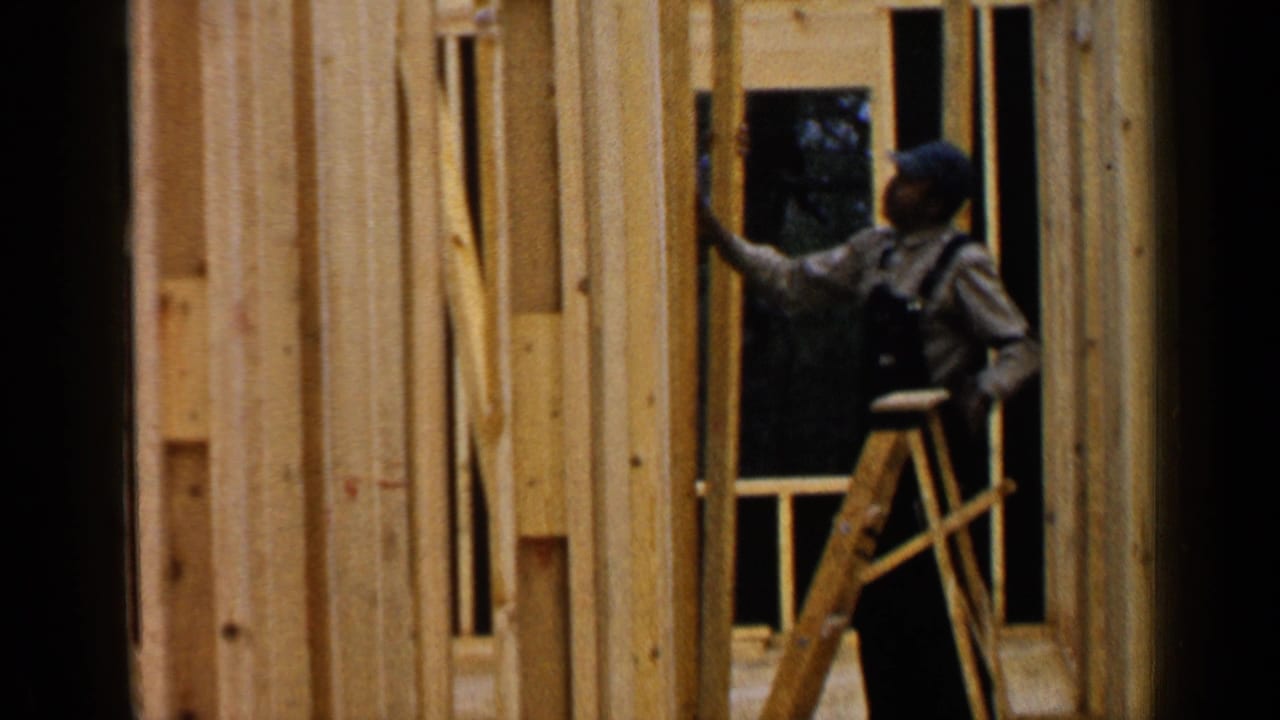 Carpenter on a ladder working beside vertical wall studs in a wood-framed structure.