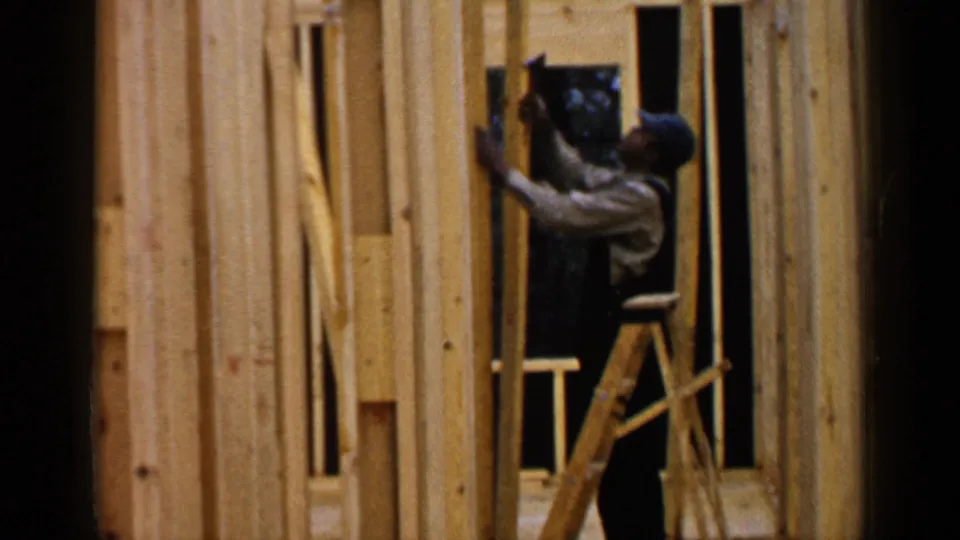 An adult carpenter in 1957 Birmingham reaches up from a ladder while working on raw wood wall framing.