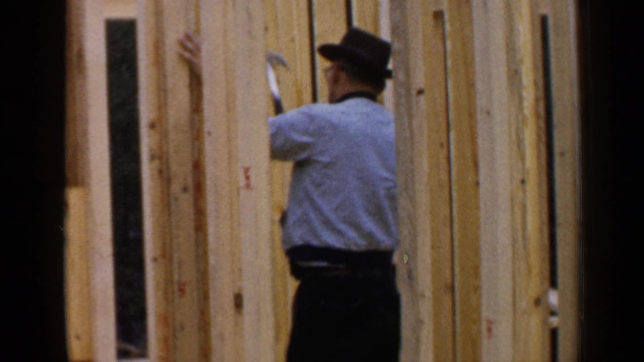 Worker in a hat stands among vertical wooden wall studs with a hand tool.